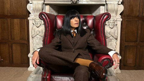 Claudia Winkleman seated in a red leather wing chair wearing a brown tweed suit, in front of a carved stone fireplace