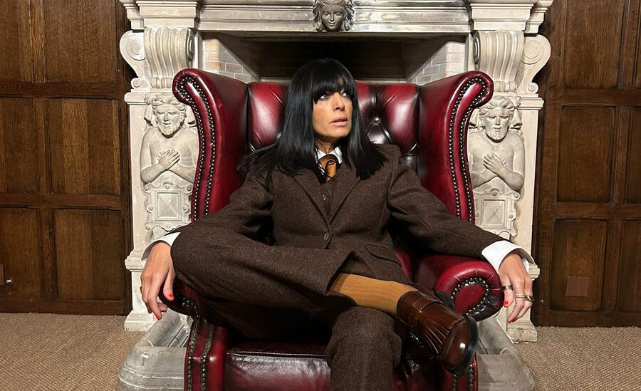 Claudia Winkleman seated in a red leather wing chair wearing a brown tweed suit, in front of a carved stone fireplace